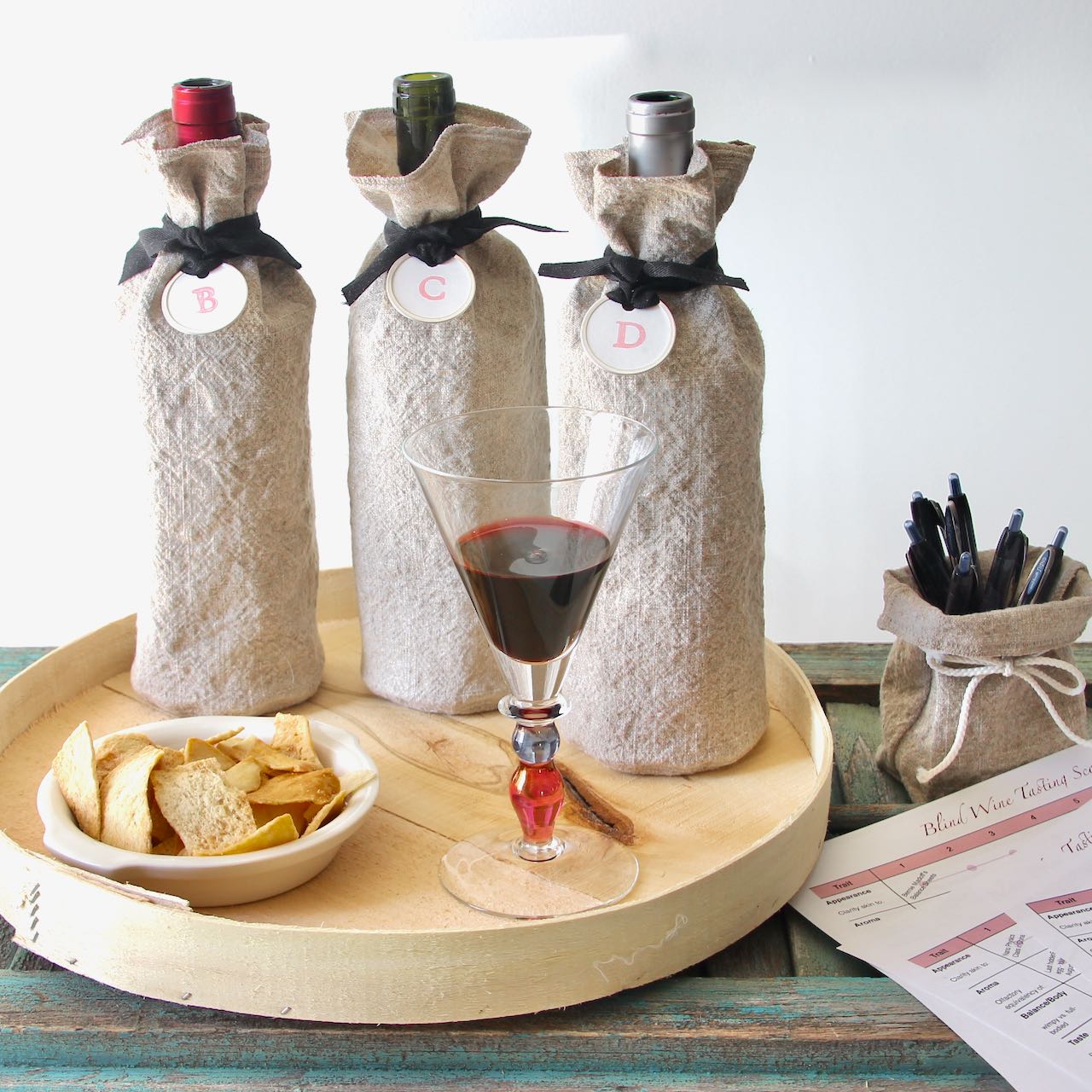 Three wine bottles in washed linen bags tied with lettered labels on a cheese box lid with a bowl of pita chips and a glass of wine poured.