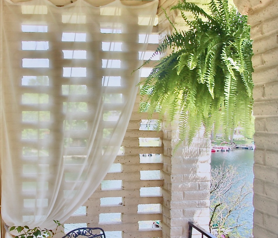A sheer curtain hanging outdoors in front of a brick screen withha Boston fern hanging beside
