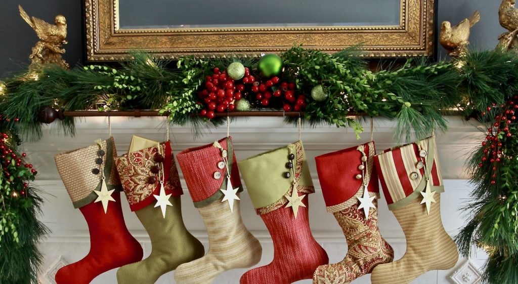 Six red green & gold Christmas Stockings hanging from a white mantel with large garland