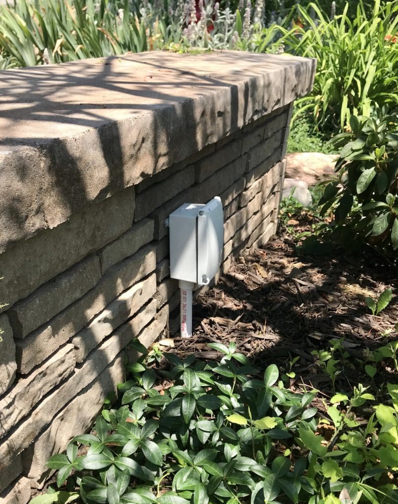 Before picture of the landscape details of white power box on pole against a stone landscaping knee wall.