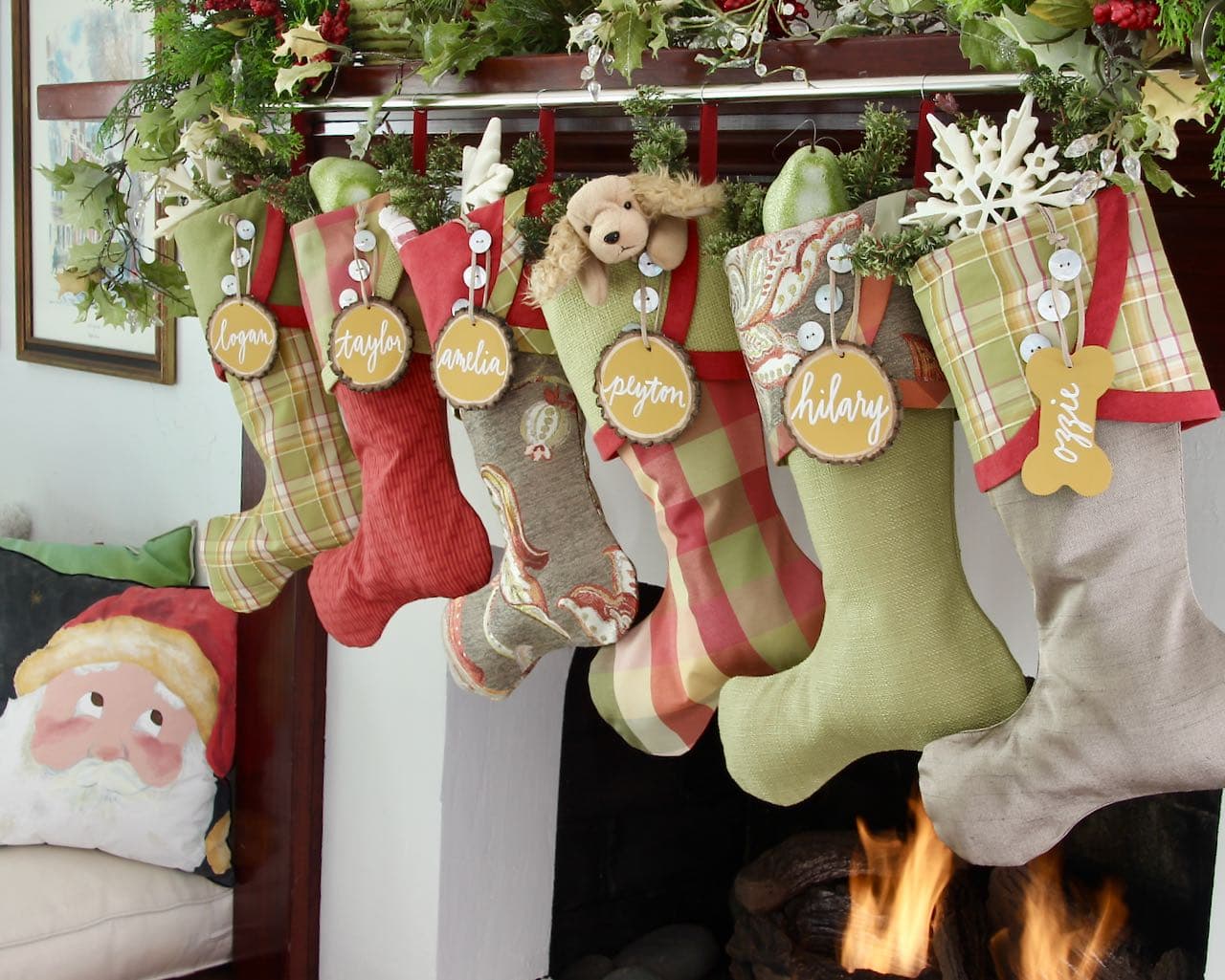 Six Christmas Stockings with Tree slice name tags viewed form a slight angle. One has a stuffed puppy peeking out of the top and there is a Santa Claus pillow next to the fire