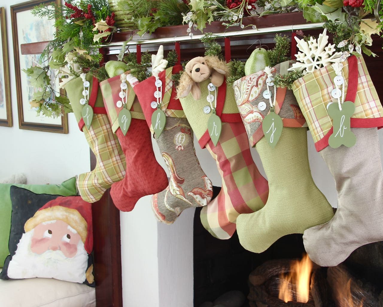 six red and green Christmas stockings with green light bulb shaped name tags hanging in front of a fire and a Santa face on a pillow is looking up at them