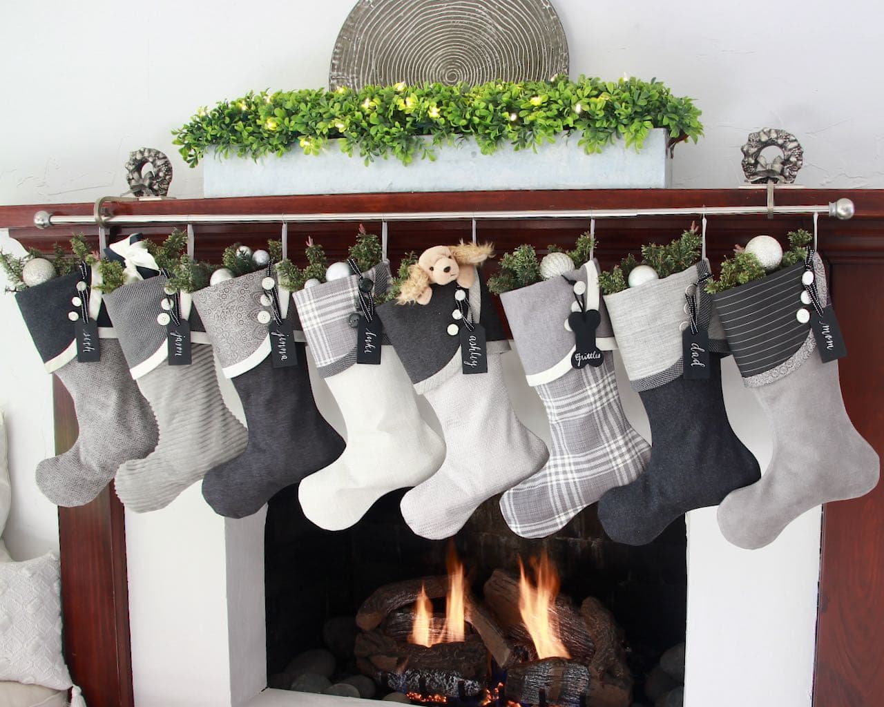 Eight coordinating Grey and winter white Christmas stockings  with black wood name tags hanging from a stocking rod on a wood mantel with a low horizontal planter filled with boxwood and a textured round art piece behind