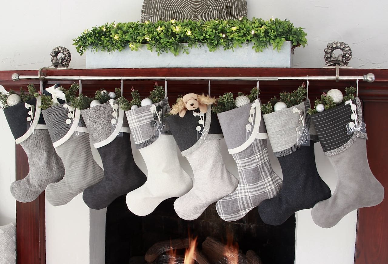 Set of eight coordinating Grey and winter white Christmas stockings hanging from a stocking rod on a wood mantel with a low horizontal planter filled with boxwood and a textured round art piece behind