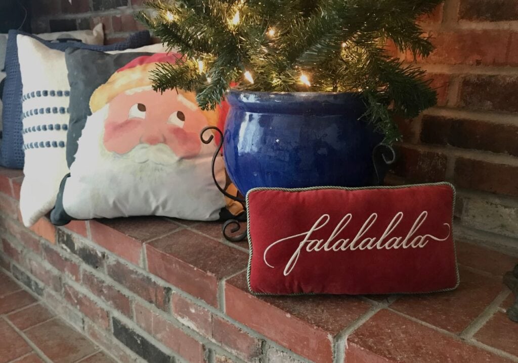 Pillow with Santa's Face looking into small tree on brick fireplace hearth with falalalala pillow