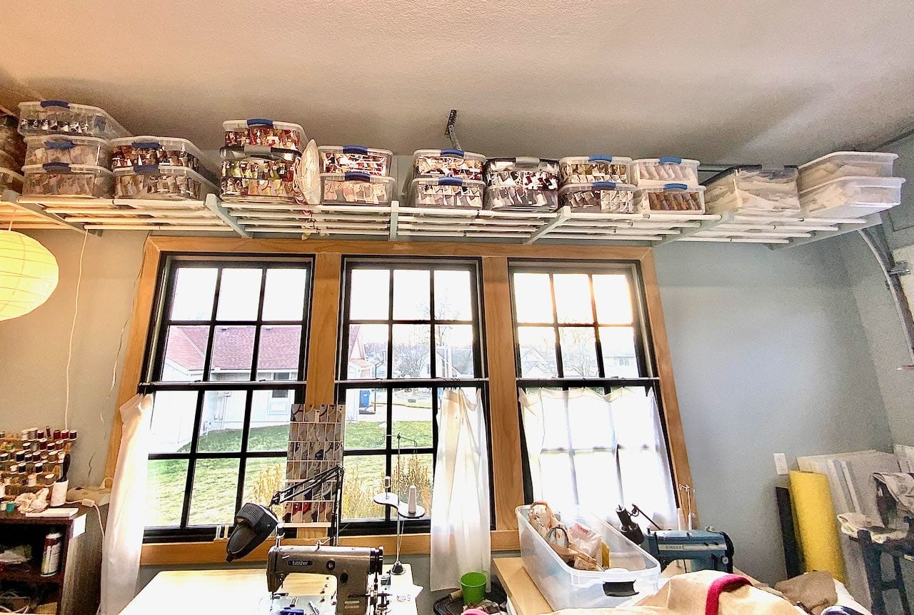 Wide view of the length of the overhead shelf filled with clear platsic tubs over bank of three large windows with sewing machines