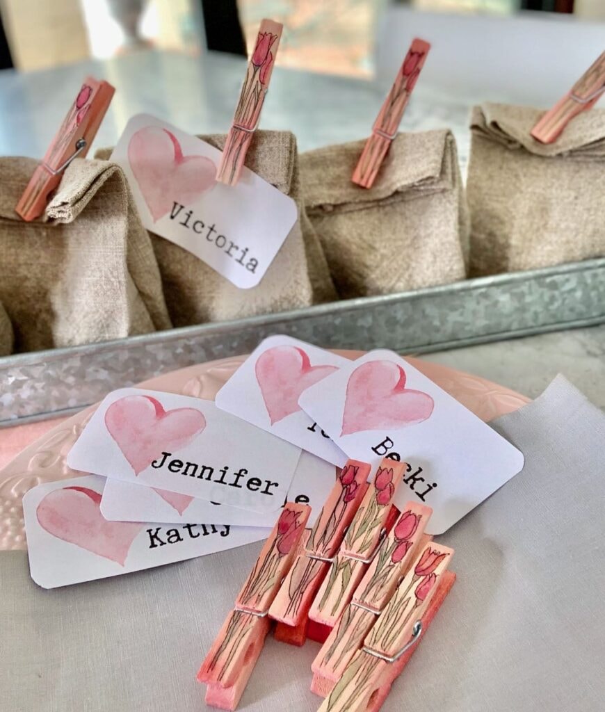 Washed linen party favor bags in a galvanized steel tray and five loose clothespins and nametags scattered on a grey and white quartz countertop