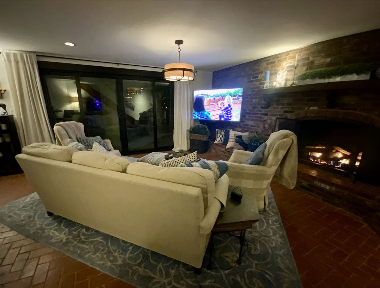 The back of a couch with two swivel wing back chairs looking at a brick fireplace wall with fire roaring and a large TV elevated in the corner and a wall of sliding glass doors at night