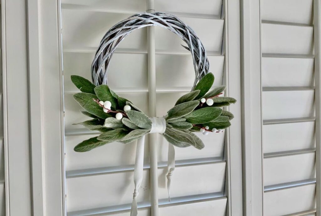 a small wood woven wreath with bundles of fresh lambs ear and faux berries is hanging on interior shutters