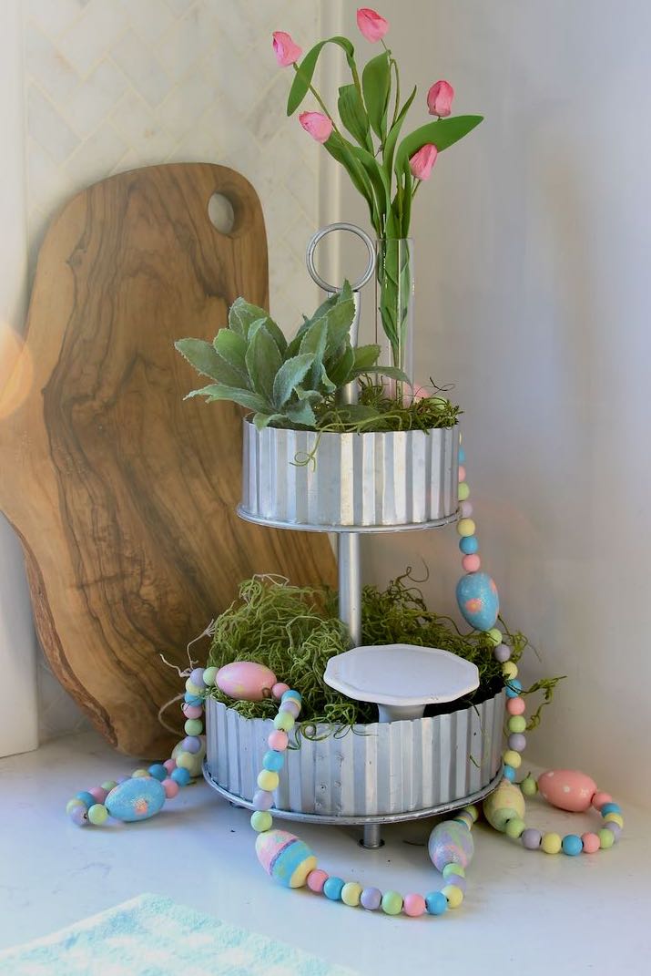 A garland of painted wooden beads and wooden eggs is on the tiered tray and pooling onto the countertop