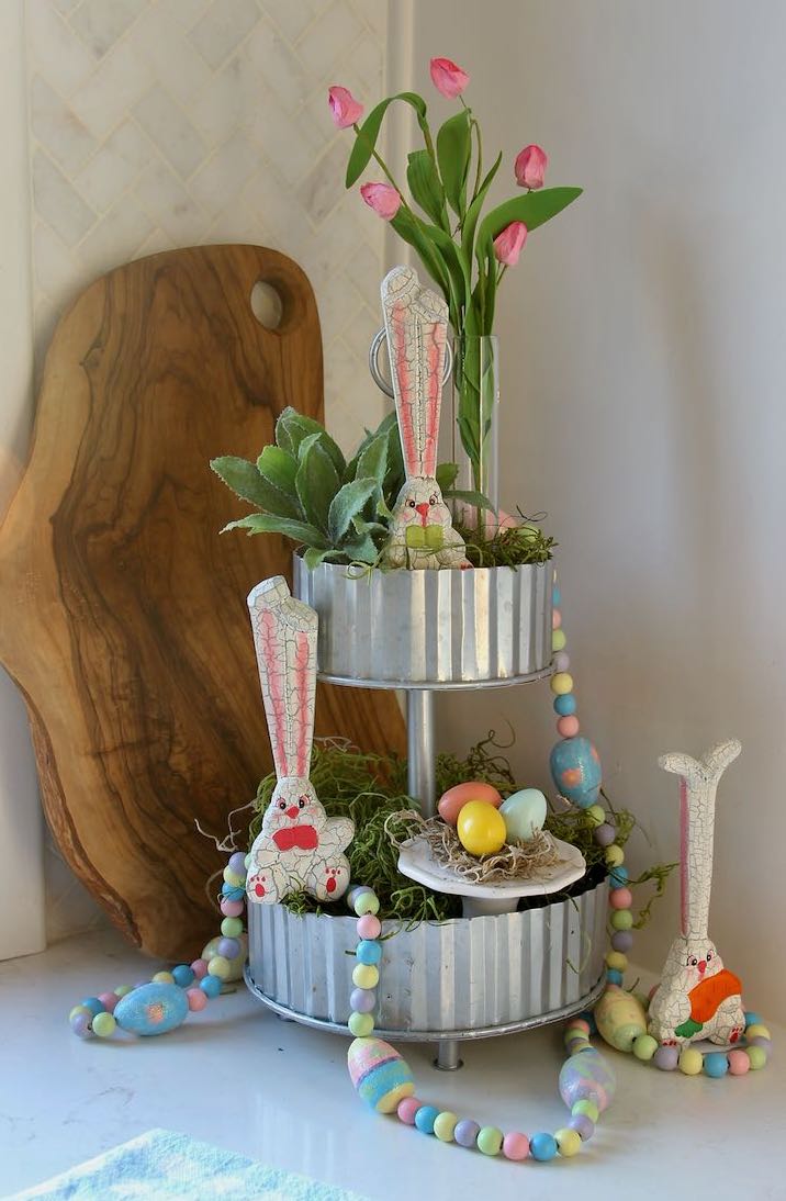 finished tiered tray with wooden Easter bunnies and garland, a nest with three painted eggsand a bud vase with Springs buds