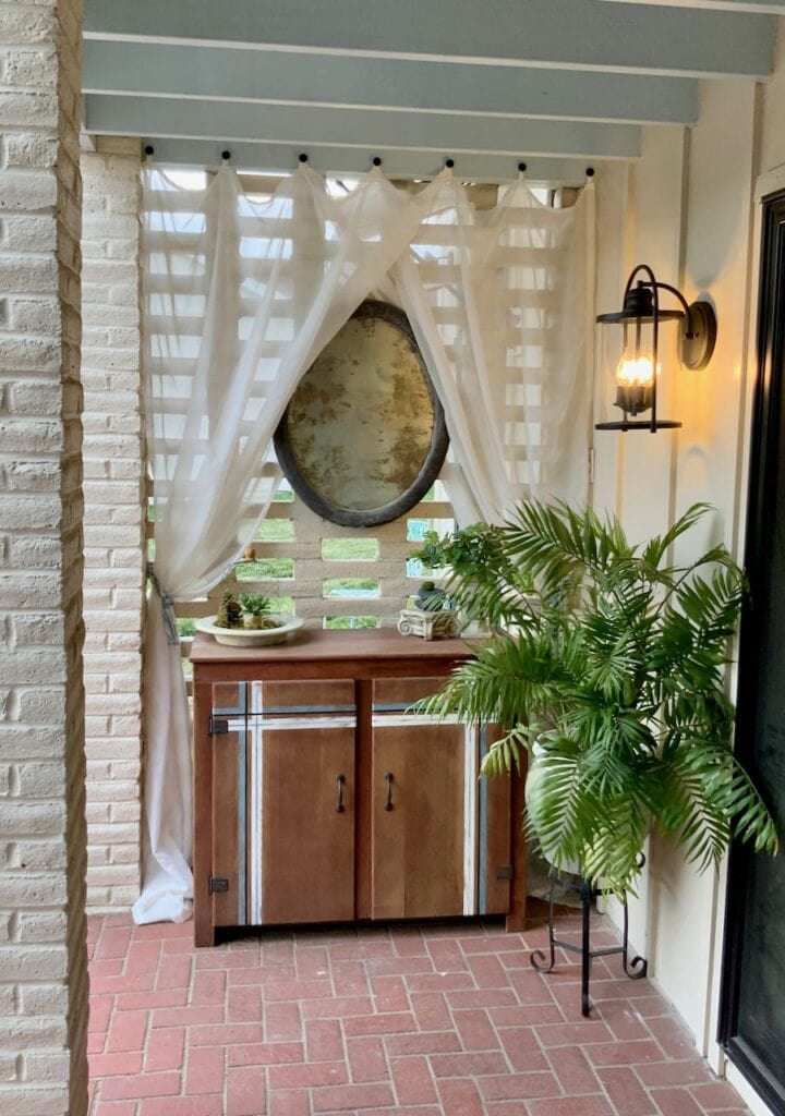 full length view of cabinet with accent striping, sheer curtains hanging on brick screen wall with an oval mirror above the cabinet and a large palm with a new light fixture