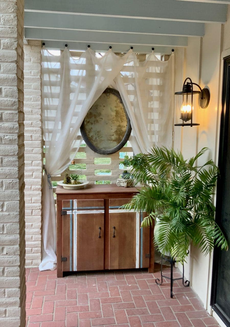 full length view of cabinet with accent striping, sheer curtains hanging on brick screen wall with an oval mirror above the cabinet and a large palm with a new light fixture