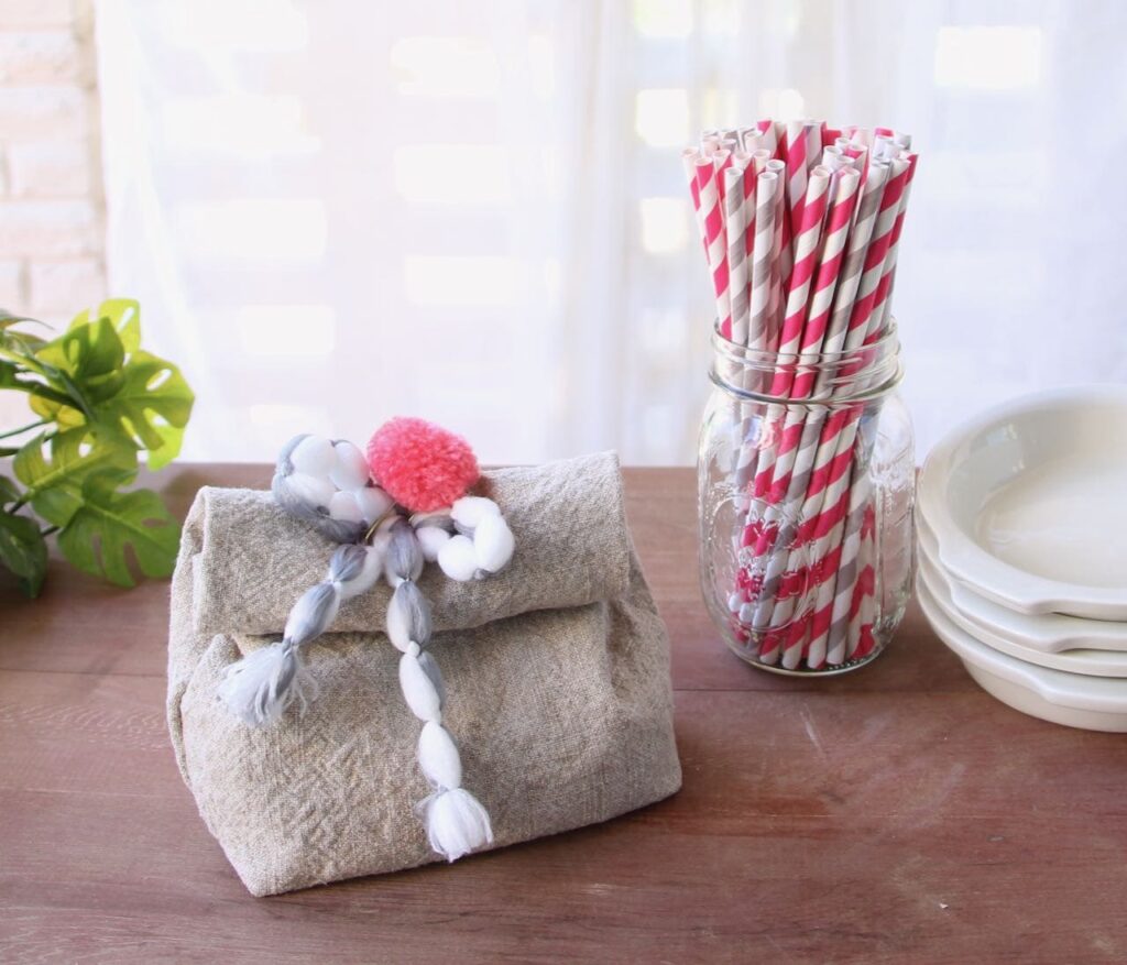 Linen Party Favor Bag with a pom pom and fluffy bow closure next to a ball jar filled with festive striped straws and a stack of snack dishes