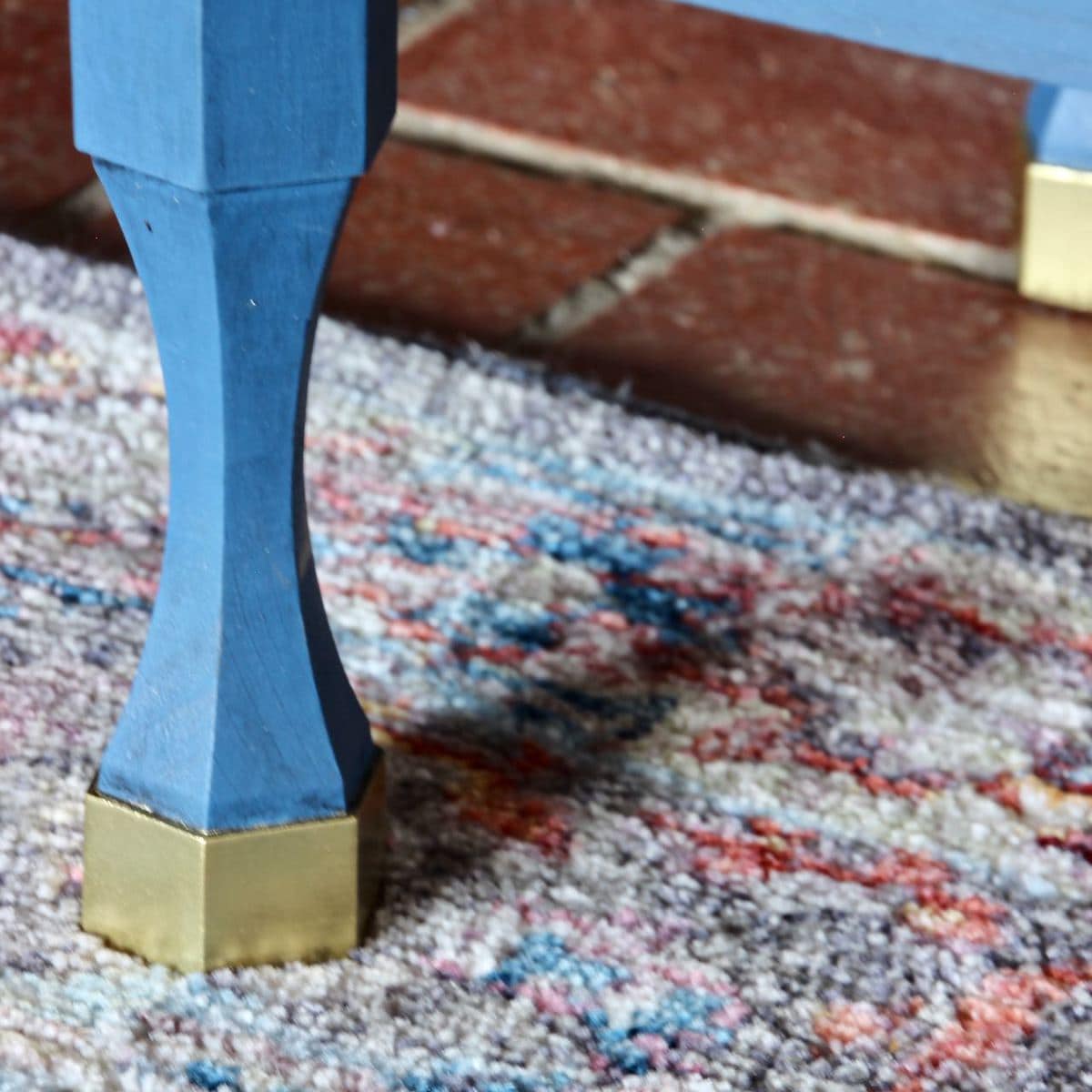 Closeup of one leg with the foot painted shiny, metallic gold sitting on a area rug with brick floor behind