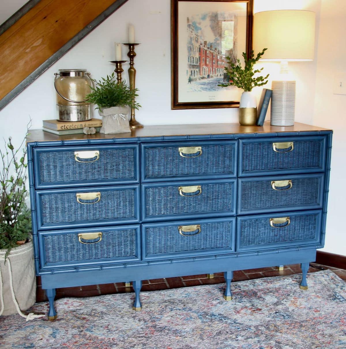 Front view of the blue chest with brass detailing, wood top and styled with a large picture, blue and white lamp, books, a green plant and a pair of burnished brass candlesticks