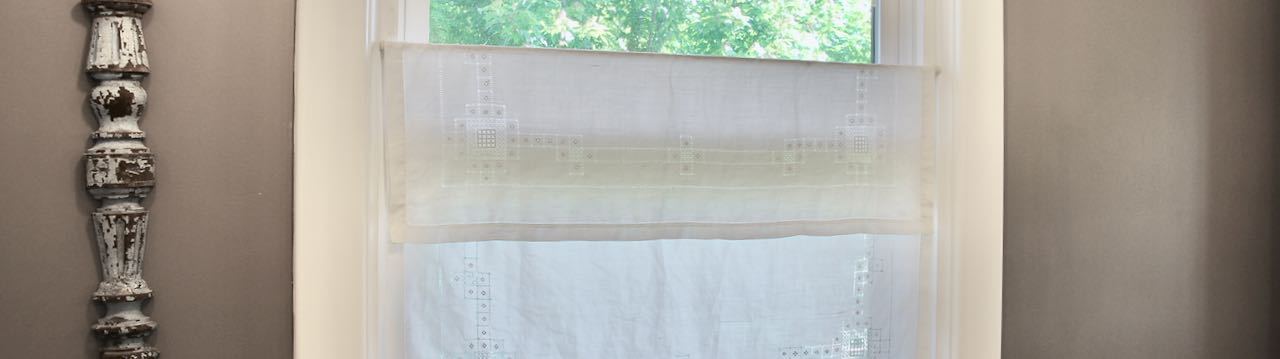 short wide view of a bathroom window with a partial view of trees and the lower portion of the window covered with a hanging vintage tablecloth over a tension rod