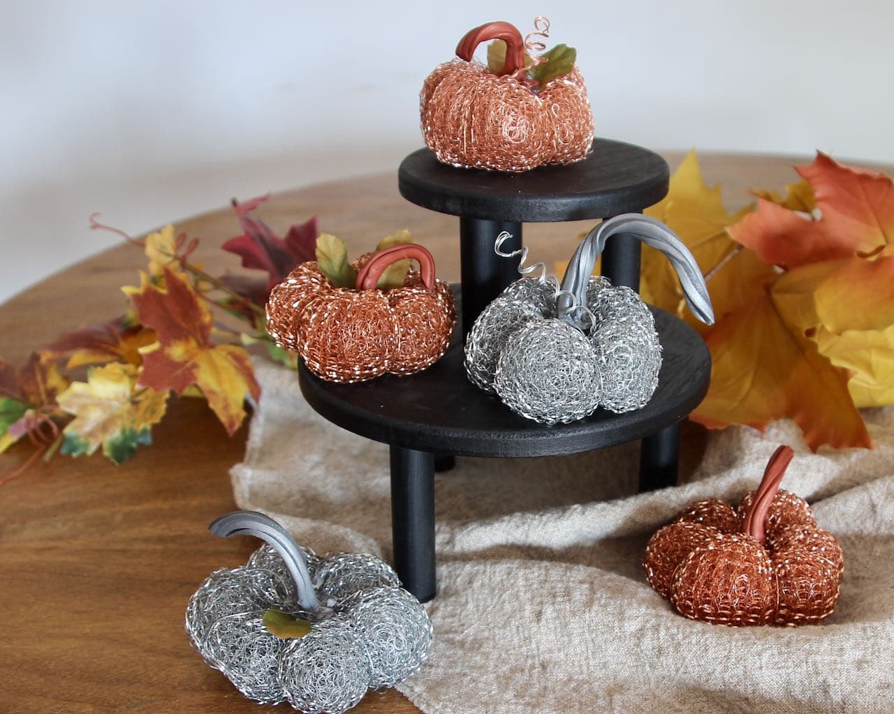 closer view of the five finished metal mini pumpkins arranged on a stack of black wood risers on a wood table with a washed linen table runner and scattered Fall leaves