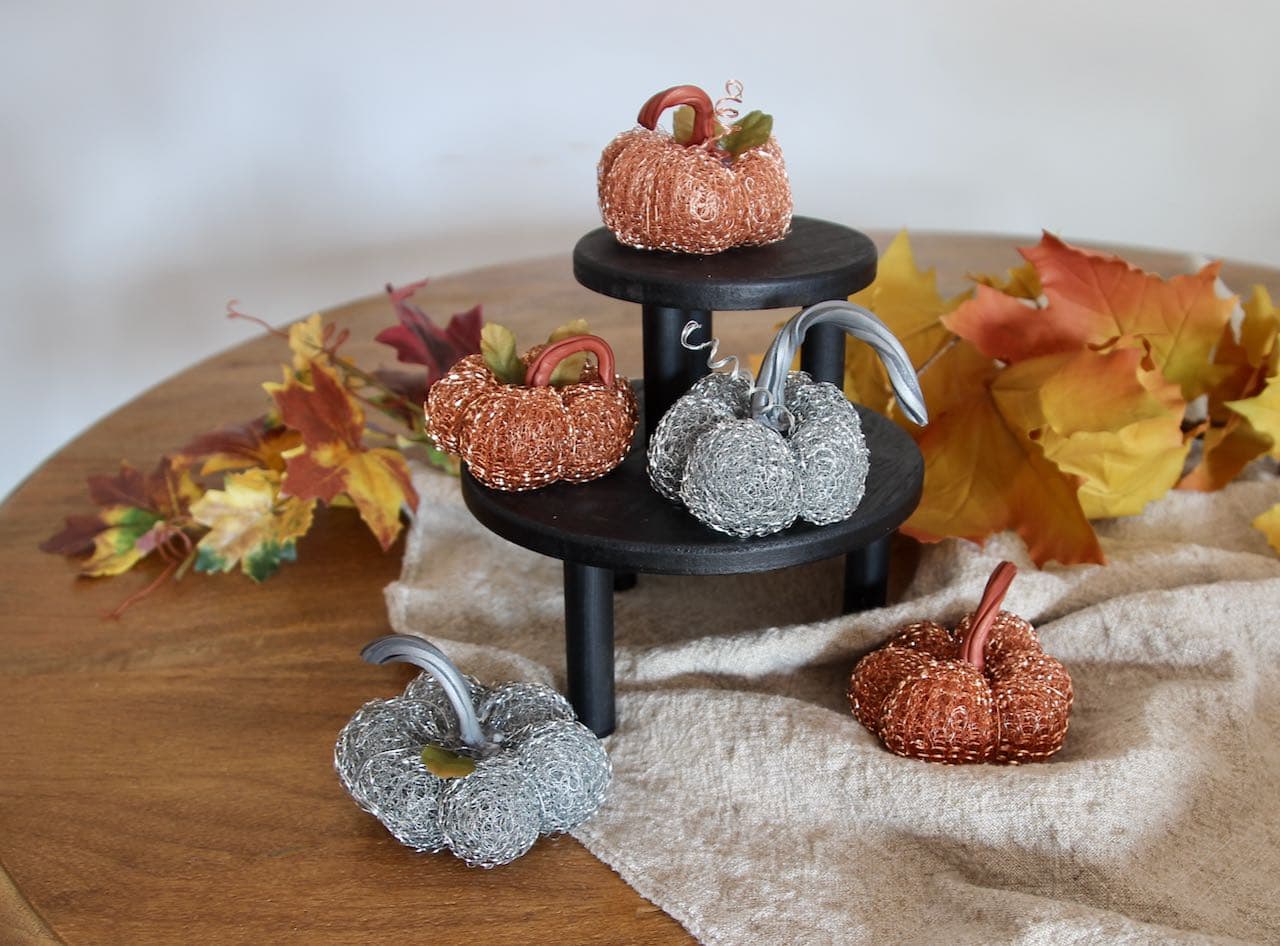 Five mini metal pumpkins, some with leaves, some with tendrils on top a stack of two black wood risers on a wood round table with a washed linen runner and Fall leaves