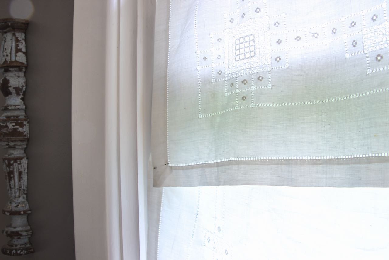 Closeup of corner of vintage table topper pinned in place over a tension rod at a bathroom window