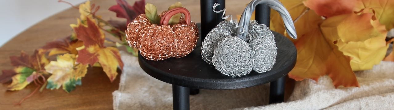 Title image of a copper and a stainless steel mini pumpkins on a black wood riser with Fall leaves scattered behind