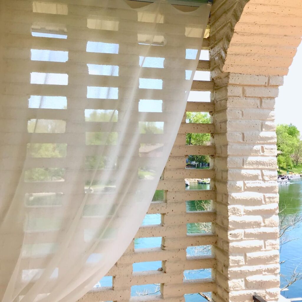 A sheer white curtain serves as a simple window treatment in front of a white brick wall with rectangular gaps, revealing a tranquil lake and lush green trees outside under a clear, sunny sky.