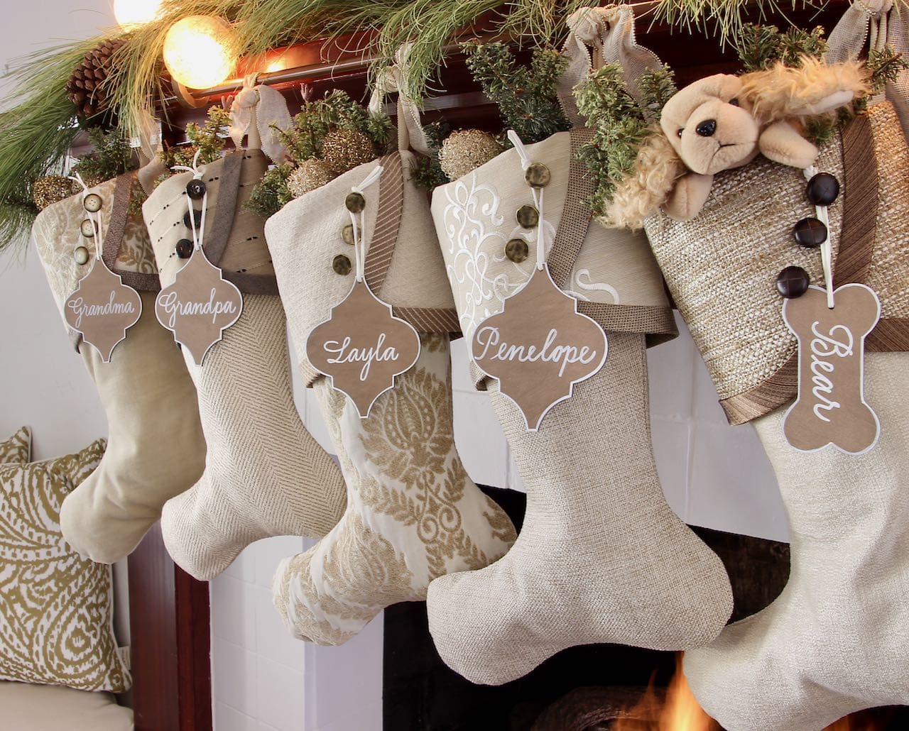 Arabesque shaped name tags are hanging on the top buttons of the stocking cuffs on 5 Warm Neutral Christmas Stockings hanging beneath an overhang of long needle pine branches