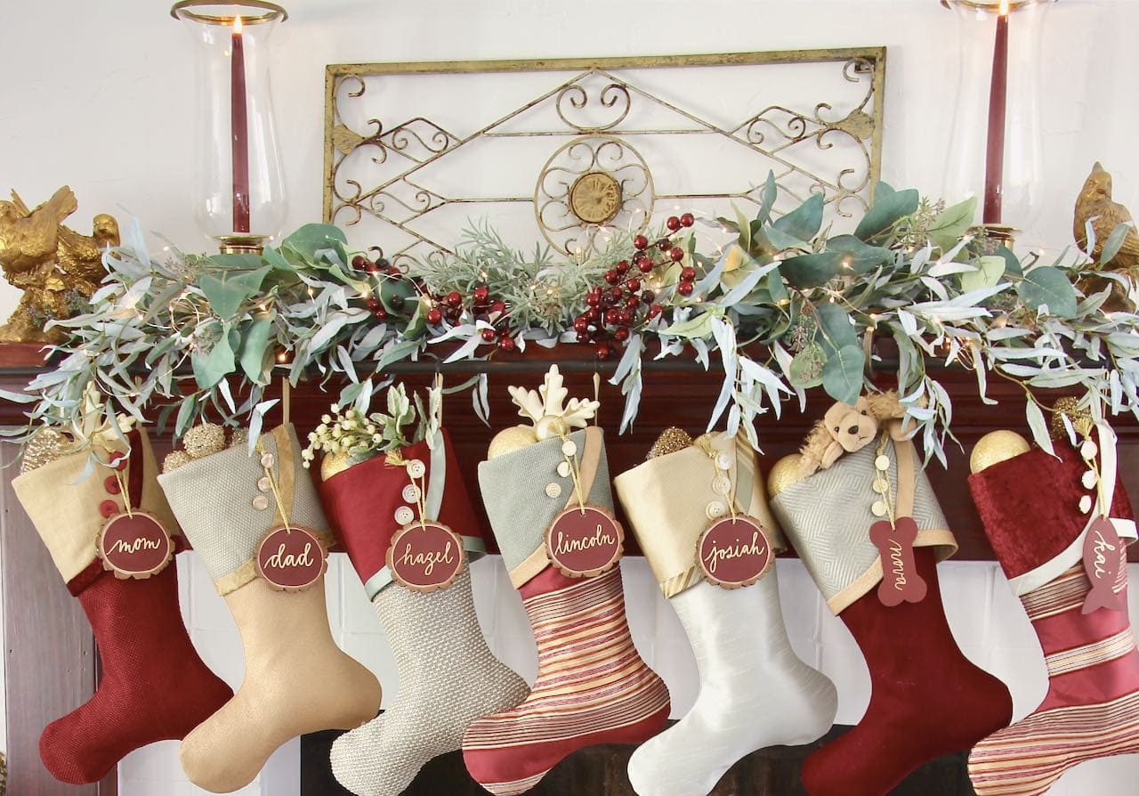 Mantel display of a vintage garden trellis on the wall flanked by a pair of glass and brass hurricane candles and seven Burgundy and Teal Christmas stocking hanging below a bough of olive branches with eucalyptus, rosemary and dark berries