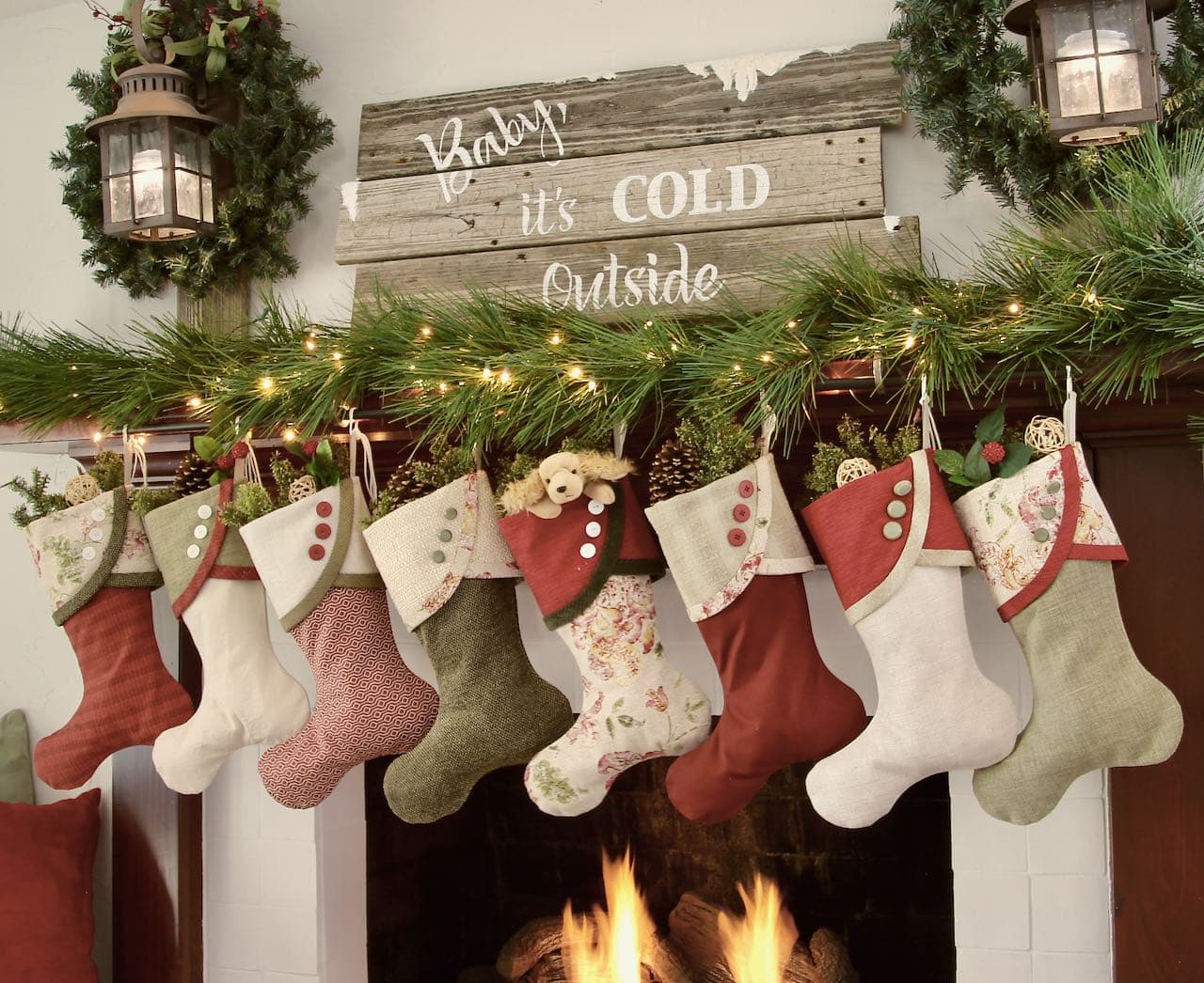 8 Cottage Christmas Stockings on a mantel with a vintage sign that reads "Baby, It's cold outside" flanked by vintage lanterns on rustic posts
