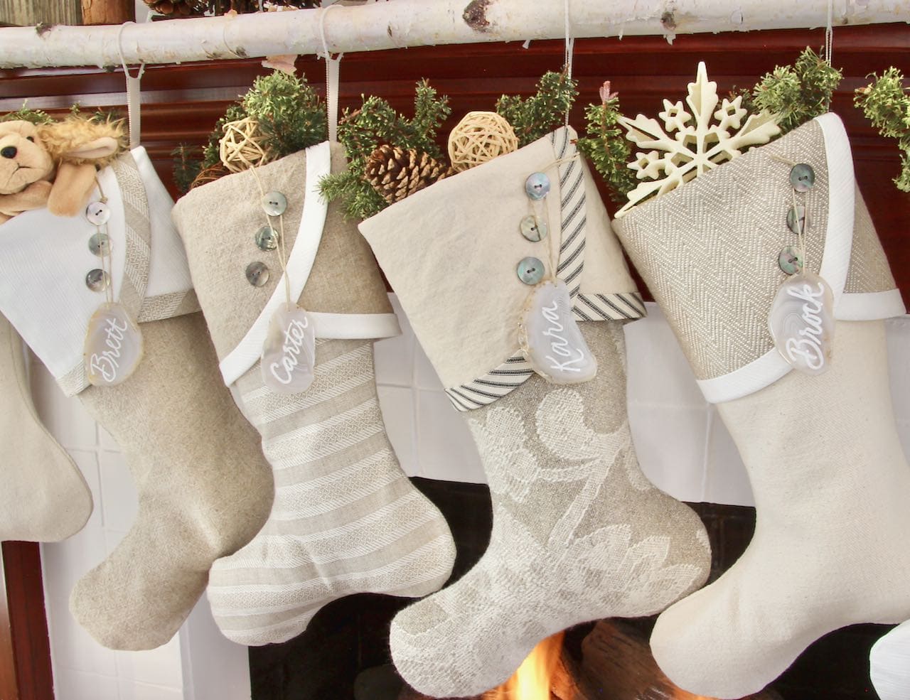 four neutral Christmas stockings with agate name tags hanging from a birch branch above a fire in the fireplace