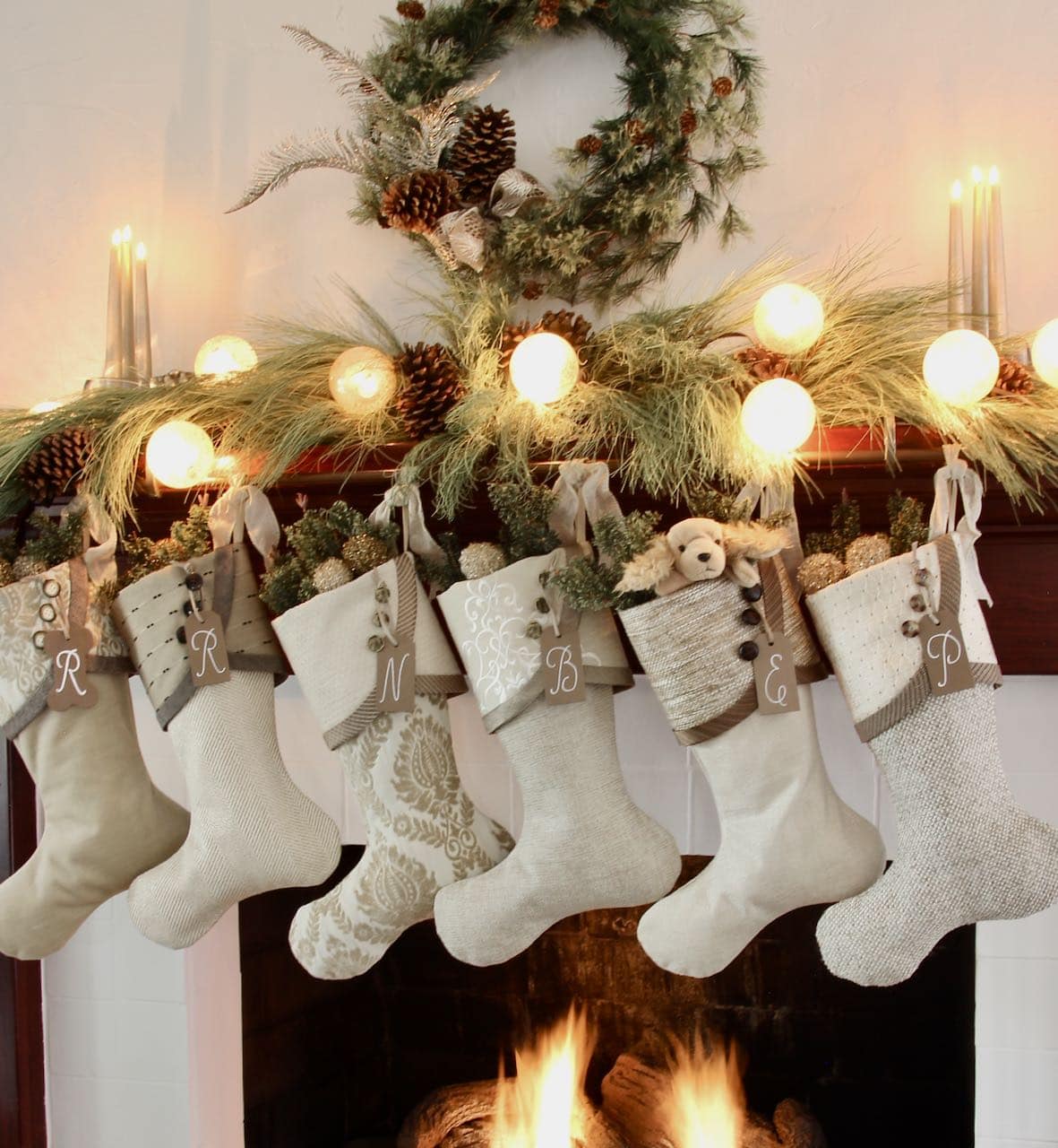 Six Warm Neutral Christmas Stockings with nutmeg initial name tags are hanging on a mantel with with simple green pine needles and globe lights, a wreath above flanked by three taper candles in s single vessel filled with sand
