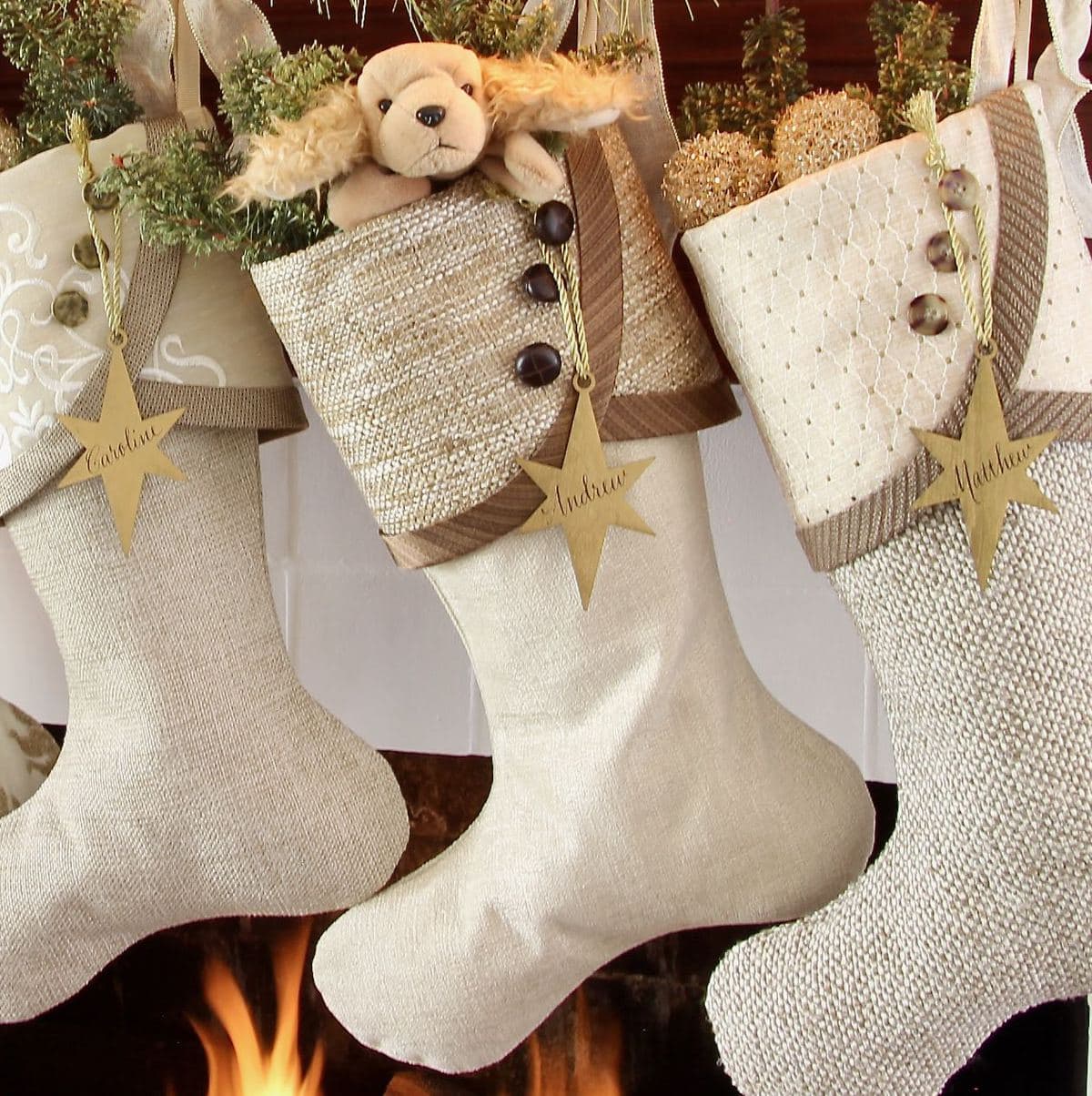 Three warm neutral Christmas stockings with gold star name tags are hanging side by side with a small stuffed puppy dog peeking out of he top of the middle one.