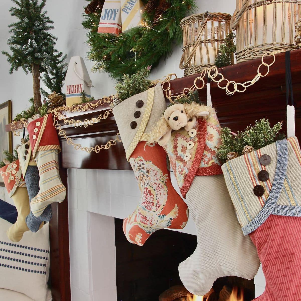 Sideview on Coastal Christmas Stockings hanging at successively lower heights, three on each side of the fireplace with wooden buoys stamped "peace", "joy" and "merry" with greens on the mantel