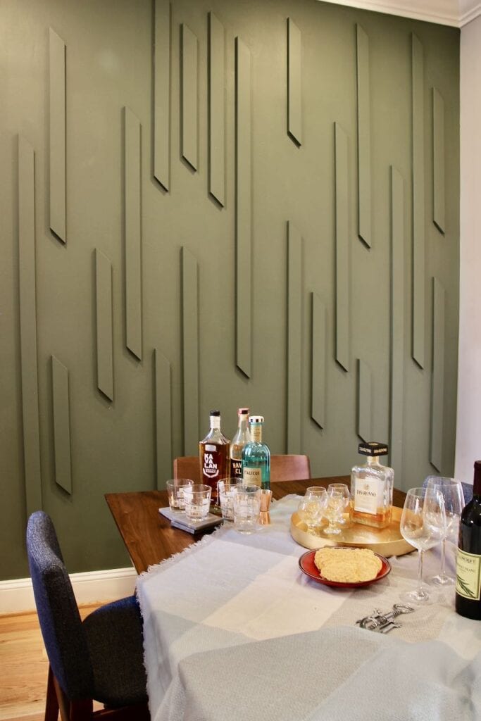 Cocktails are set out on a tablecloth at an angle on the wood table in front of the new feature wall with wood slats painted the same color as the wall