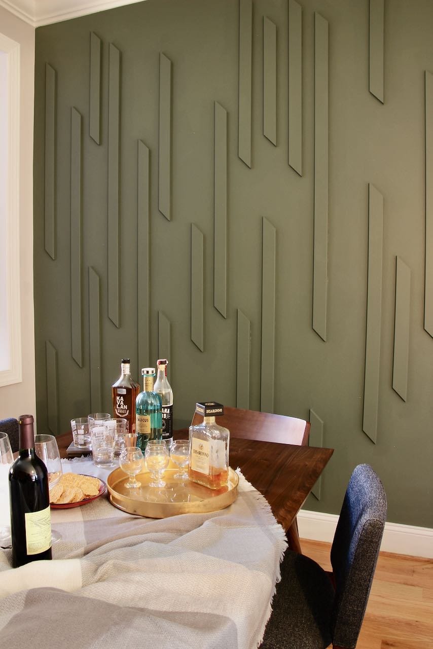 Cocktails are set out on the dark wood table with a casual tablecloth thrown at an angle in front of a modern wood slat accented dining room wall