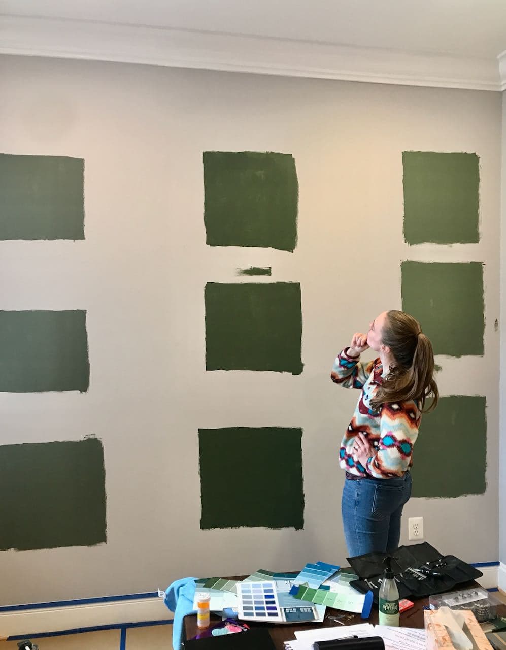 Yound adult woman with a long blonde ponytail is standing in front of a large wall that has squares of three different shades of green paint trying to decide which color she likes best