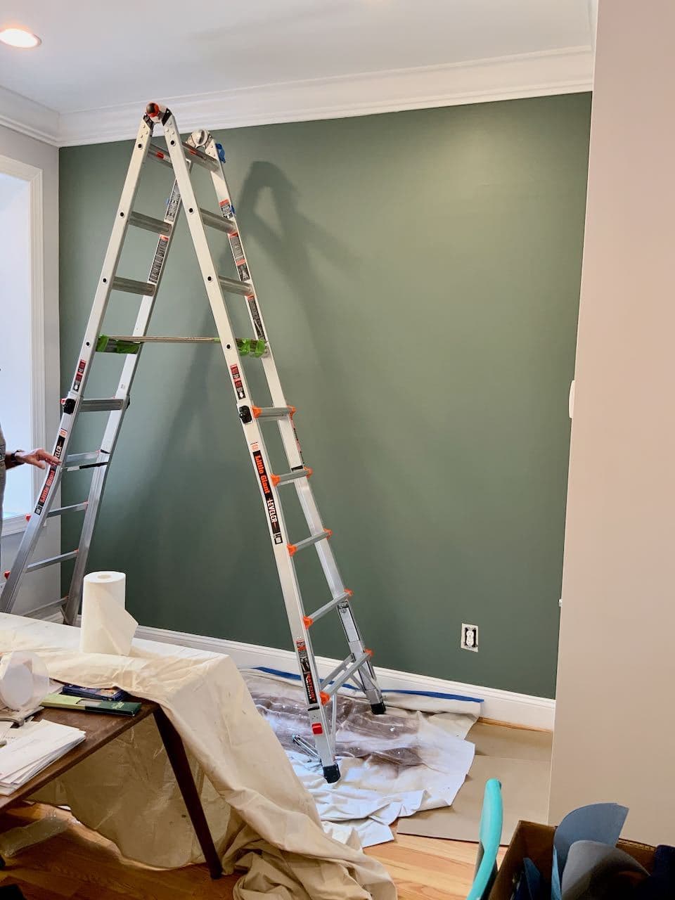 Tall ladder in front of a freshly painted smokey green wall with a drop cloth below and painters tape around