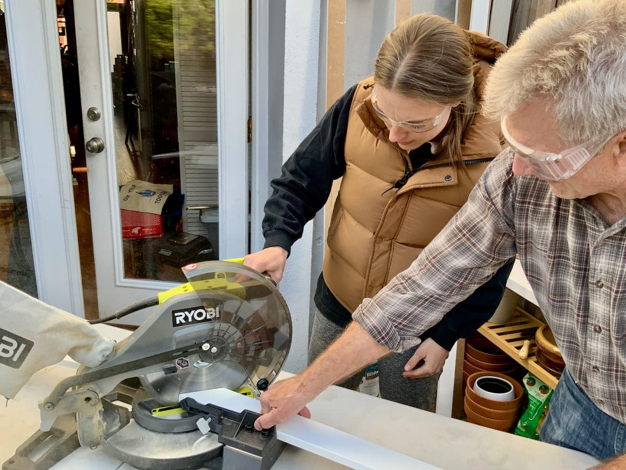 Father holding long board in a miter saw for his adult daughter to cut