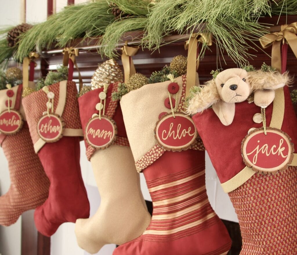 Below greenery on a mantel, four Red and Gold Christmas Stockings hanging with red tree slice name tgas hanging from the top of three buttons on each stocking cuff