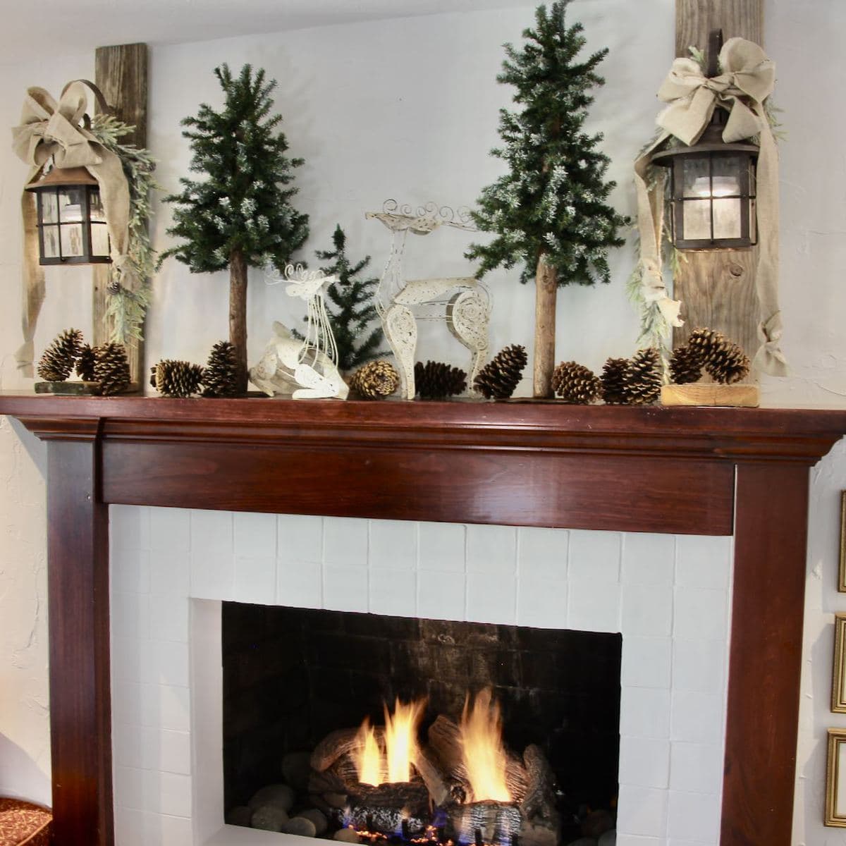 Long view of a fire in the fireplace with a wood surround and mantel with two distressed wood posts and vintage lanterns adorned with linen bows and greenery, with trees and oinecones filling the mantel between the lights