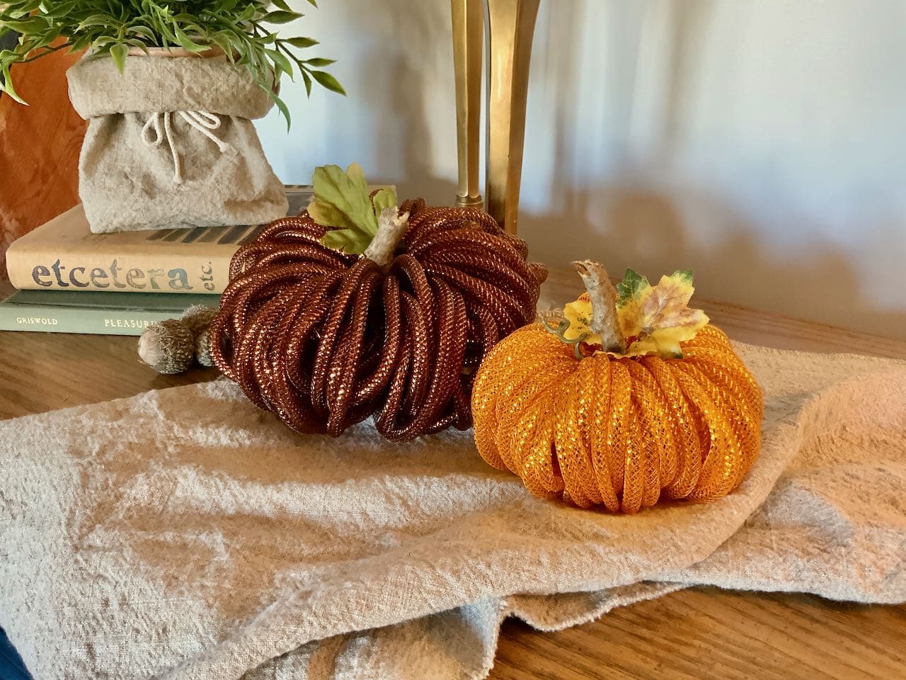 A larger bronze mesh pumpkin beside a smaller, orange mesh pumpkin on a washed linen table runner with candlestick behind