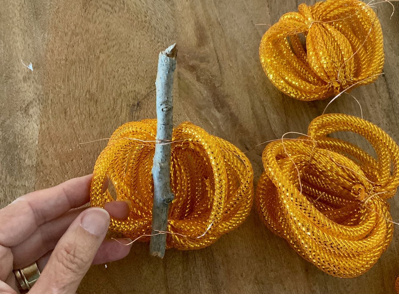 Fingers holding a stick stem with one bundle of orange mesh tubing wired to the stem at two places about 2 inches apart