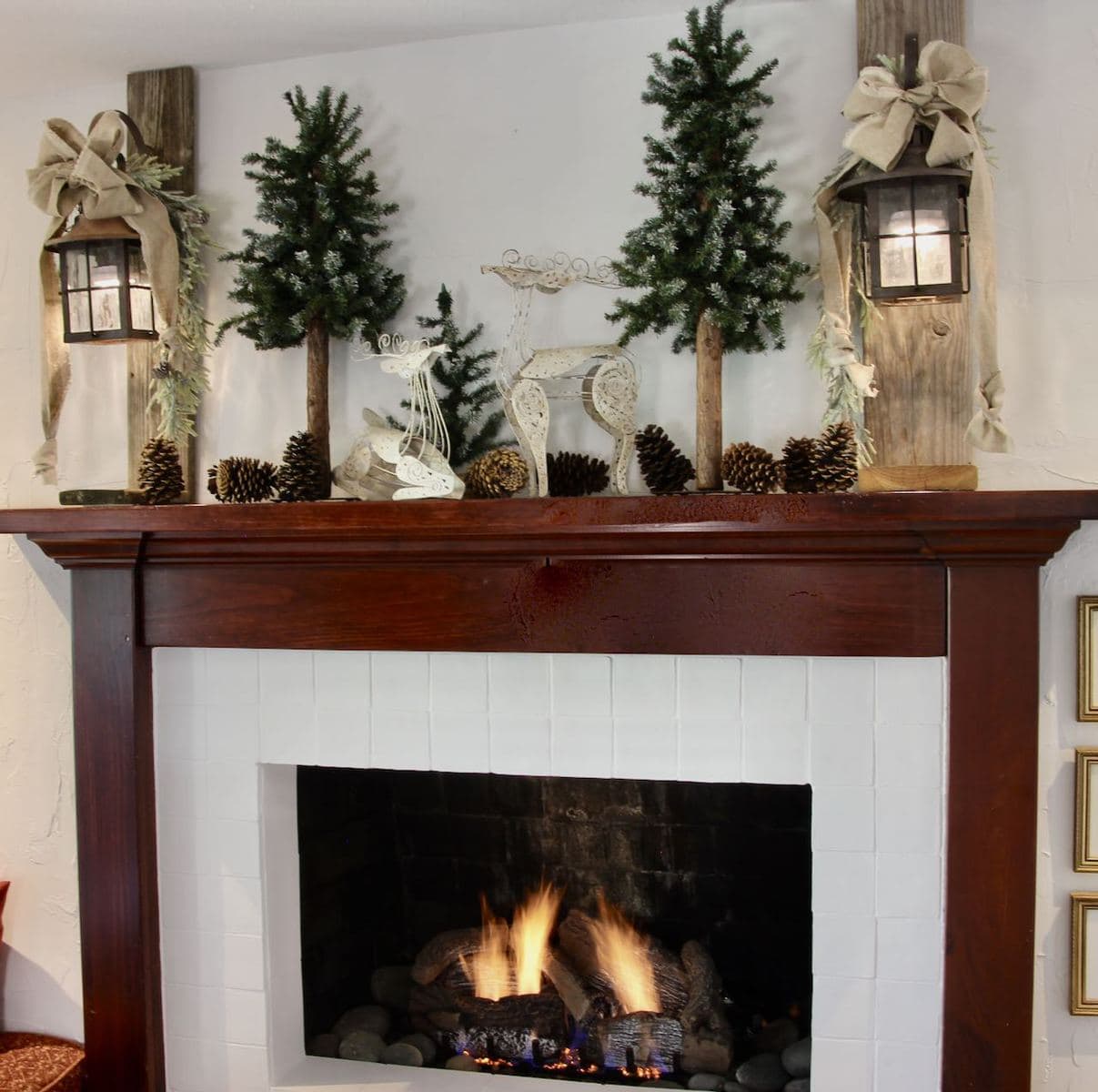 Wood Mantel above a roaring fire with a pair of distressed wood posts holding lit vintage lanterns flanking tall thin fir trees and lots of pinecones and even a pair of wire deer