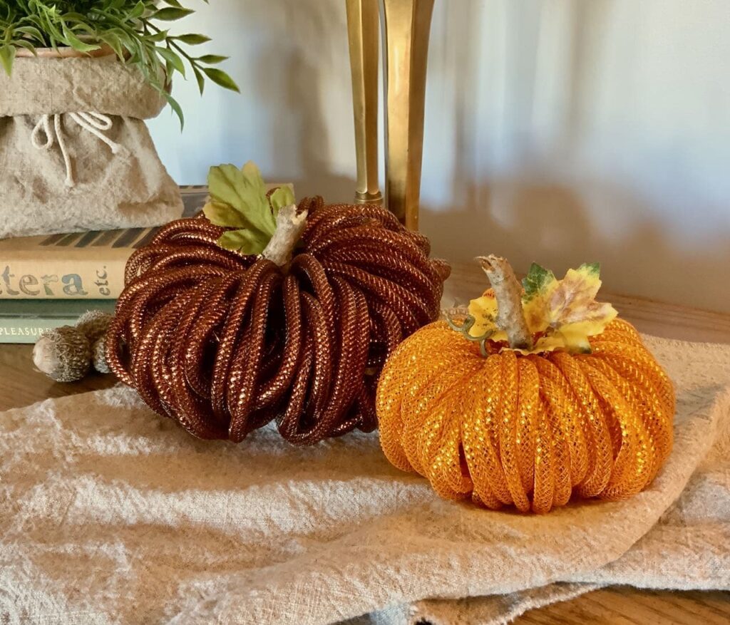 Two pumpkins made of mesh tubing are seen on a washed linen table runner with two candlesticks behind