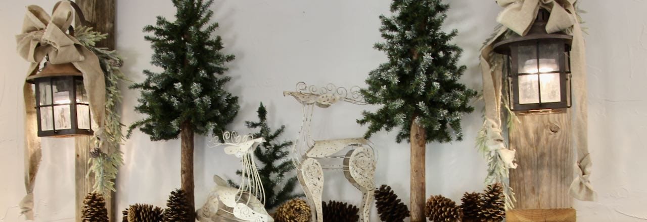 Wide view, title picture of a wood mantel   with a pair of wood posts supporting vintage lanterns that are lit and adorned with linen bows and greenery. Between the lanterns are some green trees, pinecones and a pair of wire deer