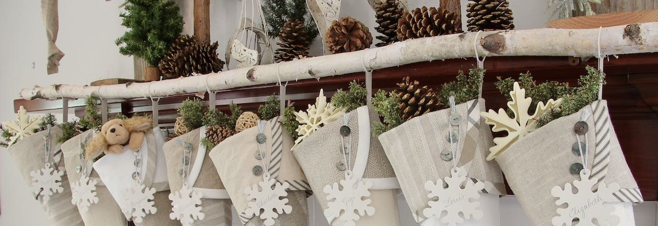 Closeup wide shot of the top half of 8 neutral farmhouse Christmas stocking with snowflake name tags hanging from a birch branch on a wood mantel