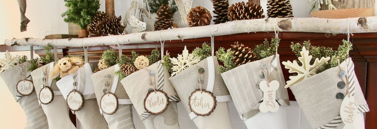 Closeup wide shot of the top half of 8 neutral farmhouse Christmas stocking with tree slice name tags hanging from a birch branch on a wood mantel