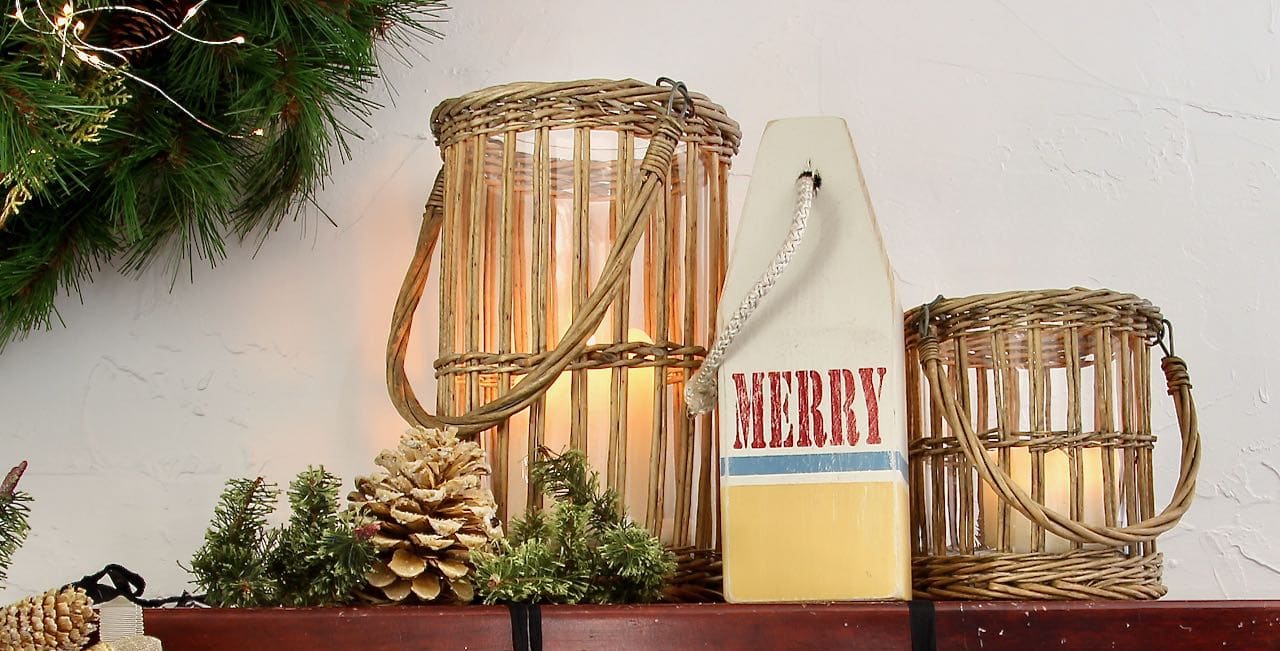 a "Merry" wooden buoy on a wood ledge flanked by candles in glass jars in rattan baskets and pinecones in front