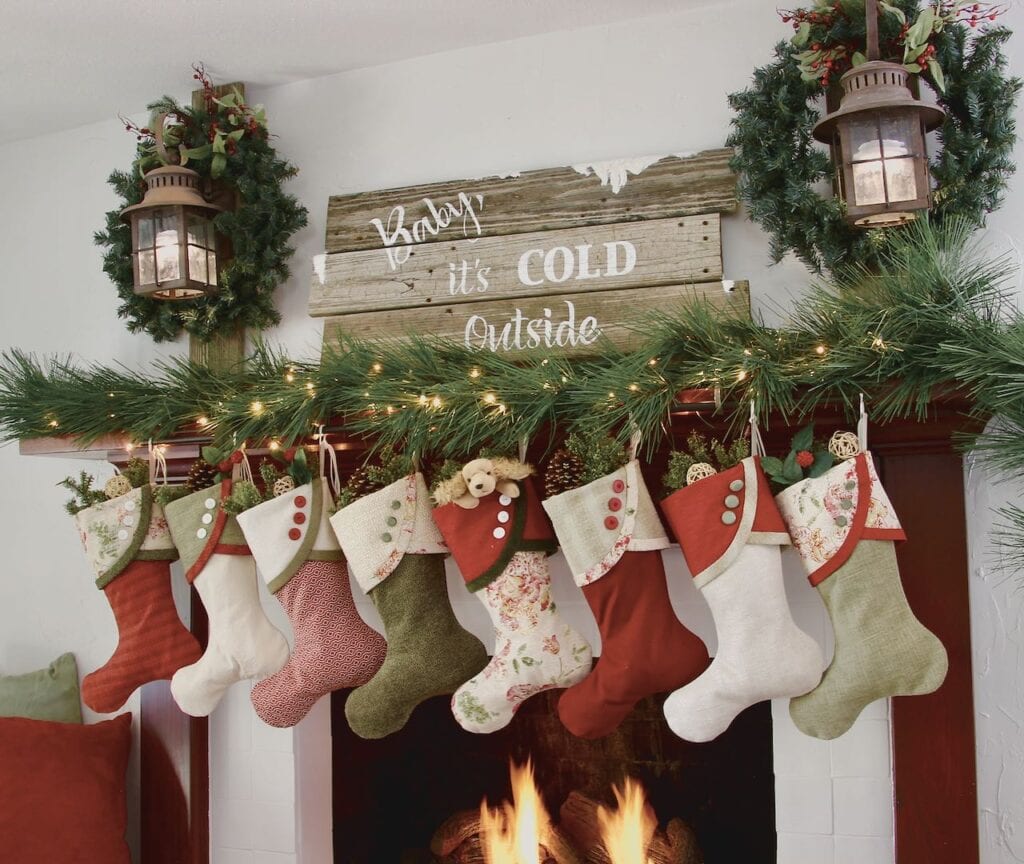 full view of fire in the fireplace with 8 Classic Cottage Christmas stockings , to lanterns above flanking a large reclaimed wood sign that reads "Baby, it's cold outside"