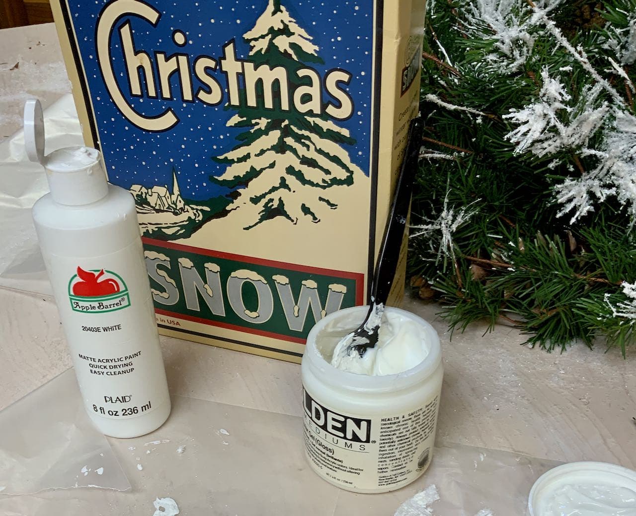 picture of a bottle of craft paint, an open jar of gel medium, a box of faux snow and an artificial fir wreath